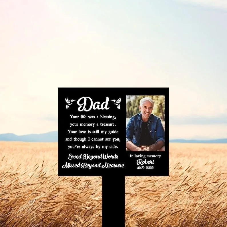 Black memorial sign in a wheat field reading “Dad” with poem lines, photo of an older smiling man and text “In loving memory Robert 1945–2022”