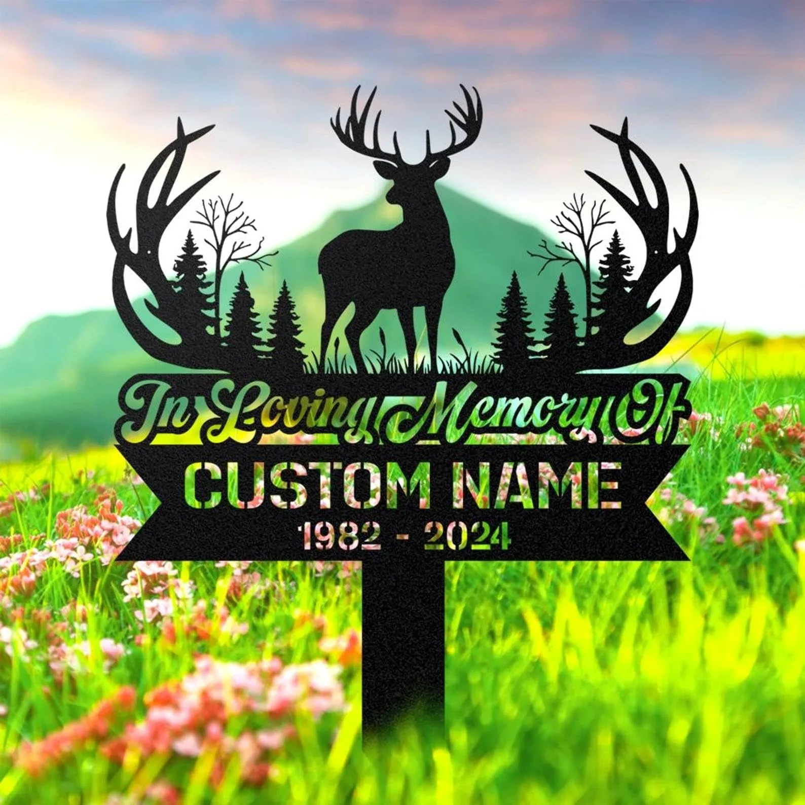 Black metal memorial sign silhouette of a stag with antlers, pine trees and antler-shaped accents reading “In Loving Memory” and placeholder “CUSTOM NAME 1982 - 2024” in a bright grassy field with flowers and distant hills.