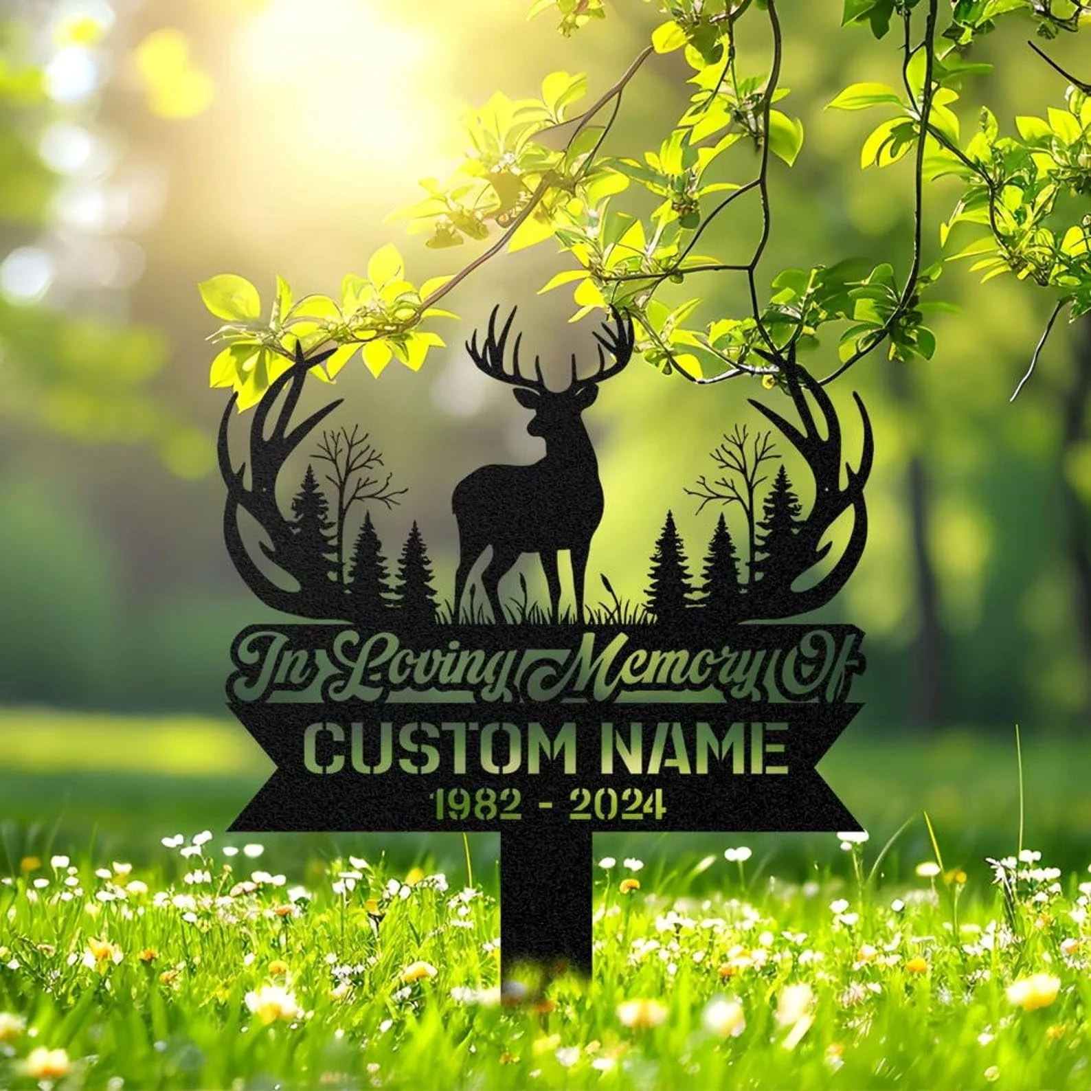 Black metal memorial lawn sign with deer silhouette, trees and antler wreath reading "In Loving Memory" and placeholder text "CUSTOM NAME 1982 - 2024" in a sunny grassy garden