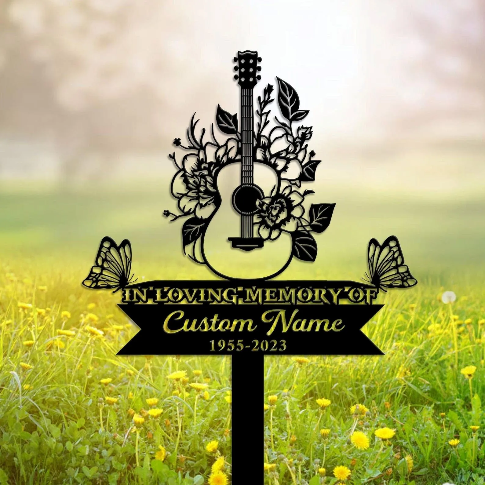 Black memorial sign in a grassy field featuring a floral acoustic guitar silhouette, butterflies and text reading "In Loving Memory" with placeholder "Custom Name 1955-2023"