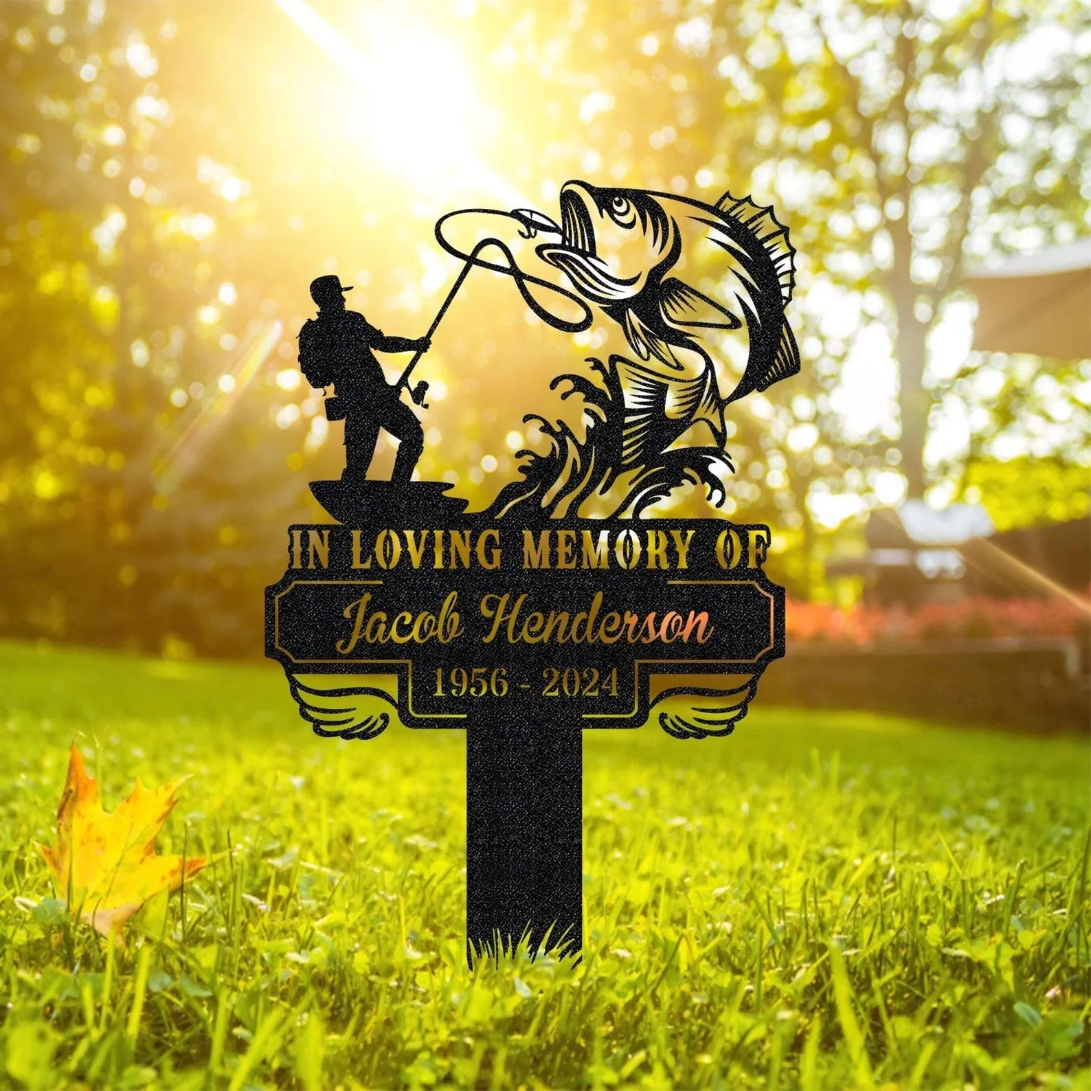 Black memorial yard sign showing a fisherman catching a large fish with text "In Loving Memory of Jacob Henderson 1956 - 2024" staked in sunny grassy yard