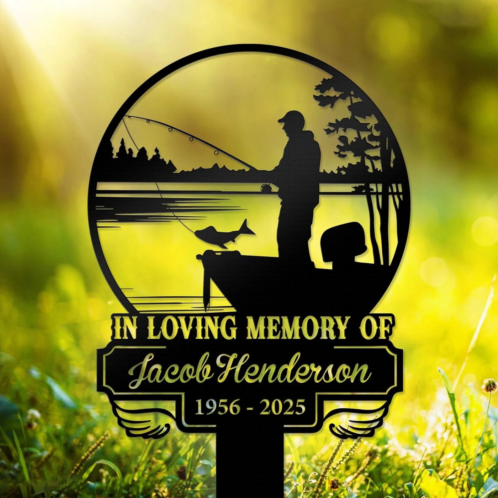 Black silhouette memorial sign showing a man fishing from a boat with a fish on the line, framed in a circular lakeside scene and text reading "In Loving Memory of Jacob Henderson 1956 - 2025" against a sunlit grassy background