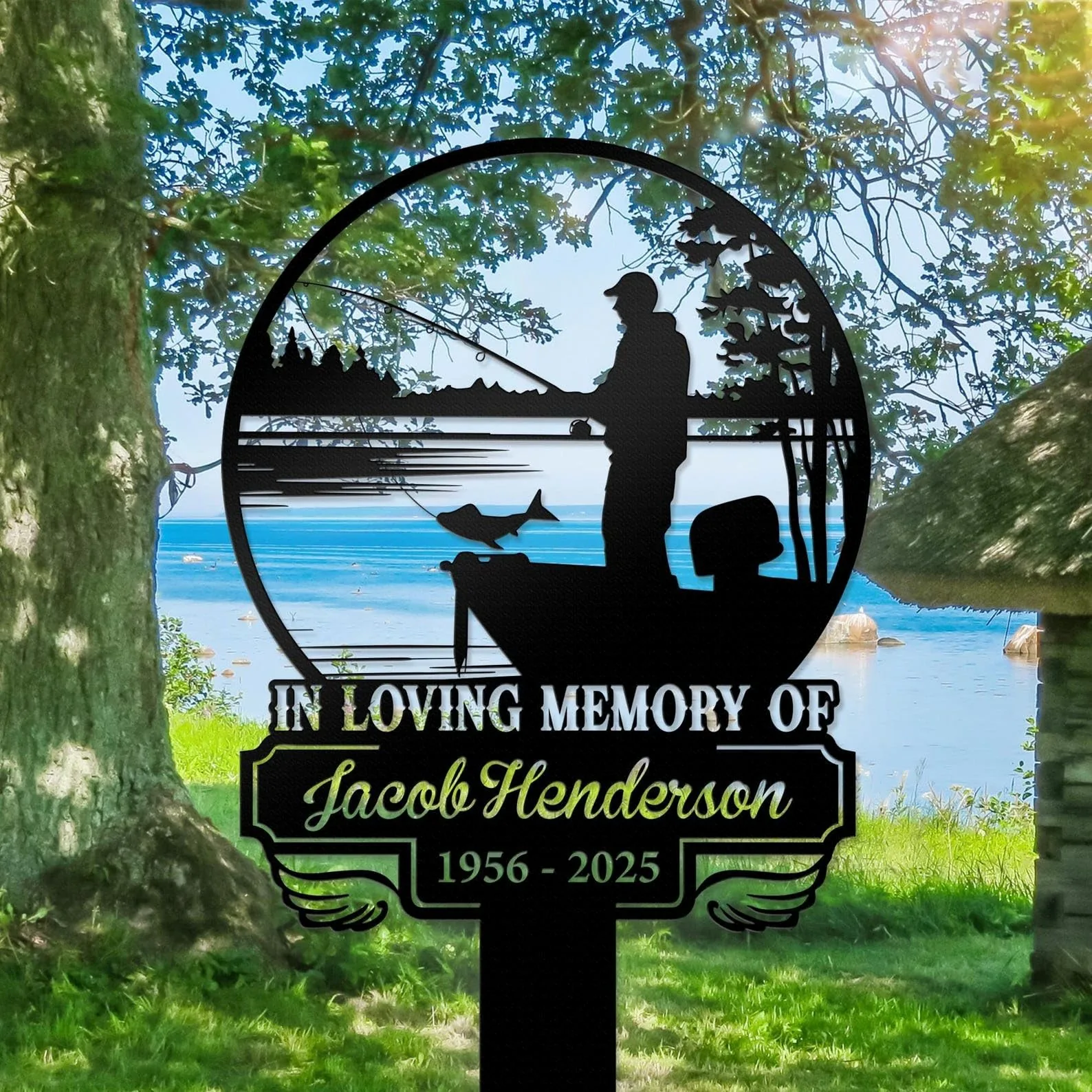 Black memorial yard sign with a fisherman silhouette and fish over a lakeshore scene, text reading “In Loving Memory of Jacob Henderson 1956 - 2025,” placed on grassy lakeside under trees