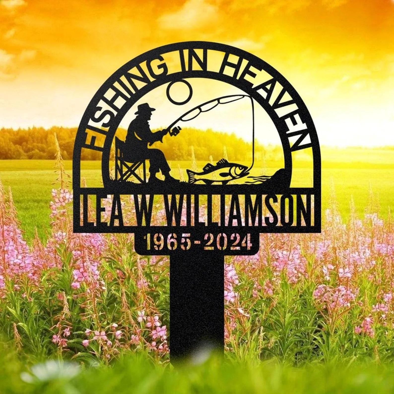 Black metal memorial sign reading "Fishing in Heaven" with silhouette of a seated fisherman and a fish, personalized name and years, shown in a flower field at sunrise