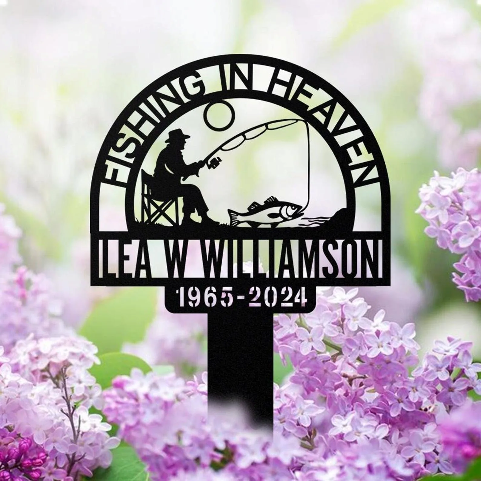 Black metal memorial garden sign with silhouette of a man fishing, text “FISHING IN HEAVEN,” name “ILEA W WILLIAMSON” and dates “1965-2024,” set against blurred lilac flowers
