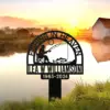 Metal memorial sign reading "Fishing in Heaven" with silhouette of fisherman and fish, name "Ilea W Williamson" and dates "1965-2024" placed by a lakeside at sunrise