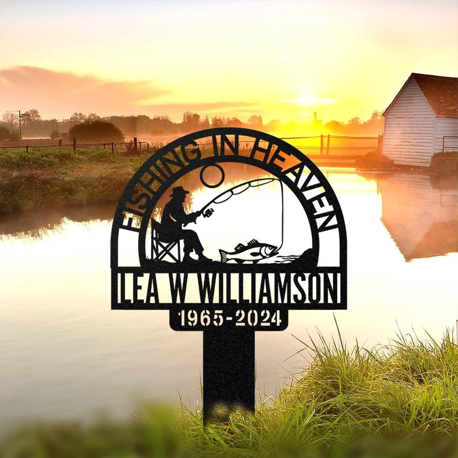 Metal memorial sign reading "Fishing in Heaven" with silhouette of fisherman and fish, name "Ilea W Williamson" and dates "1965-2024" placed by a lakeside at sunrise