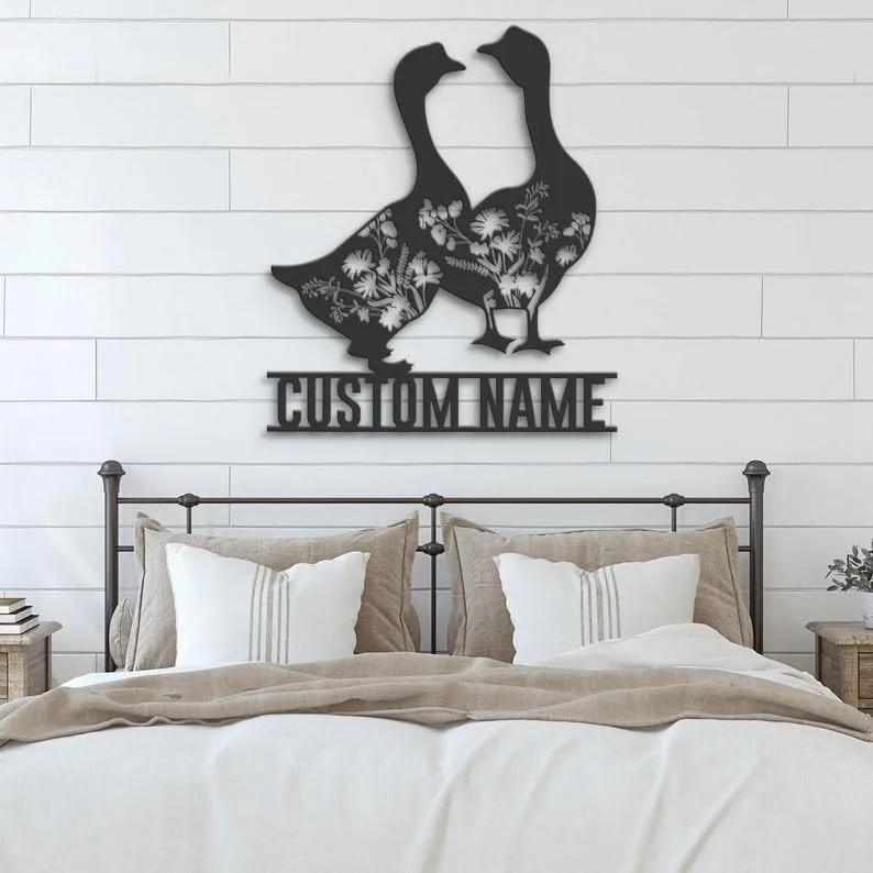 Black metal wall art of two geese with floral cutouts and the words "CUSTOM NAME" mounted above a farmhouse bed with neutral pillows and shiplap wall