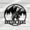 Round metal wall art showing two cyclists kissing on bikes in front of pine trees and mountains with placeholder text “CUSTOM NAME” mounted on a white wooden wall