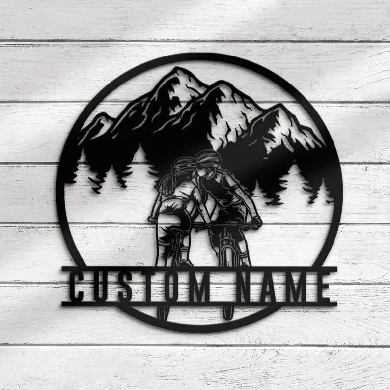 Round metal wall art showing two cyclists kissing on bikes in front of pine trees and mountains with placeholder text “CUSTOM NAME” mounted on a white wooden wall