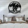 Metal wall art of two cyclists riding in front of pine trees and mountains with placeholder text "CUSTOM NAME" mounted above a farmhouse bed
