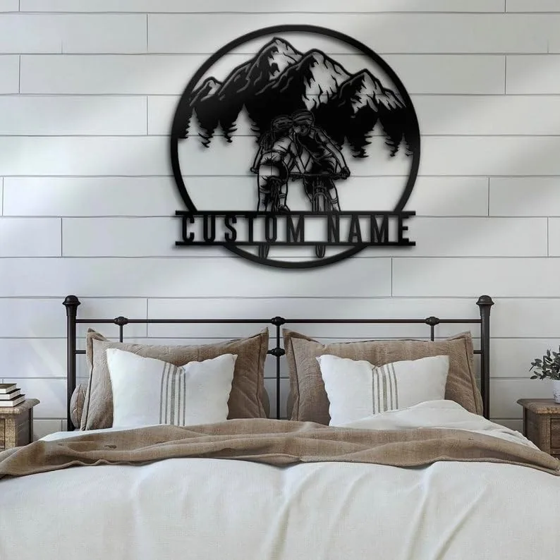 Metal wall art of two cyclists riding in front of pine trees and mountains with placeholder text "CUSTOM NAME" mounted above a farmhouse bed