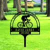 Black metal yard memorial sign with a cyclist silhouette and the name "David James" and years "1967-2024" set in a green park with trees and flowerbeds