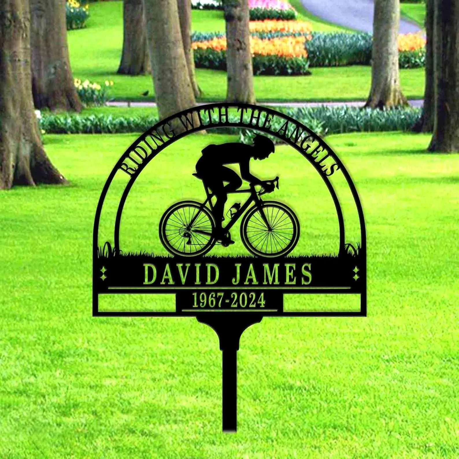 Black metal yard memorial sign with a cyclist silhouette and the name "David James" and years "1967-2024" set in a green park with trees and flowerbeds