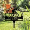 Black metal garden memorial sign with a cyclist silhouette and text “DAVID JAMES 1967-2024” staked in a sunny grassy yard with plants and an orange chair in the background