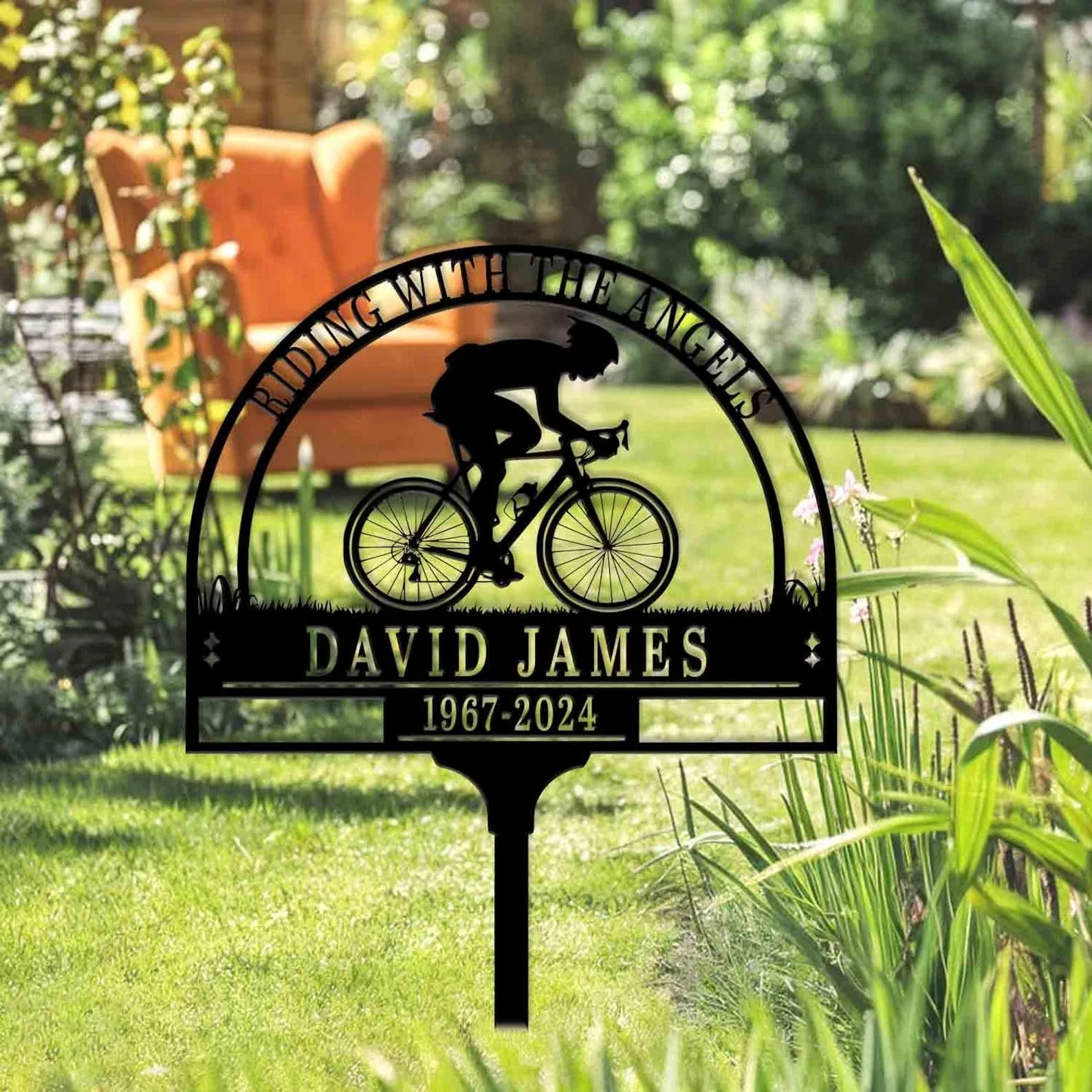 Black metal garden memorial sign with a cyclist silhouette and text “DAVID JAMES 1967-2024” staked in a sunny grassy yard with plants and an orange chair in the background
