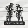 Black metal wall art of two motocross riders holding hands riding toward viewer with a rectangular plate below reading “CUSTOM NAME,” mounted on white wood background