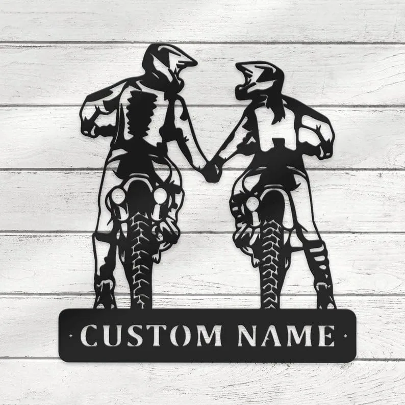 Black metal wall art of two motocross riders holding hands riding toward viewer with a rectangular plate below reading “CUSTOM NAME,” mounted on white wood background