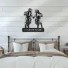 Black metal wall art of two motocross riders holding hands above a customizable nameplate, hung on white shiplap wall over a neutral-toned bed with pillows and bedside tables