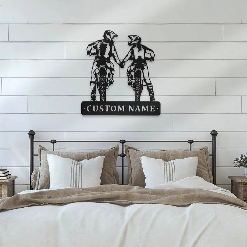 Black metal wall art of two motocross riders holding hands above a customizable nameplate, hung on white shiplap wall over a neutral-toned bed with pillows and bedside tables