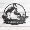 style-8.-Flamingo-Couple-1 Black metal wall art of two flamingos in a circular frame with tropical leaves and a ribbon reading 'CUSTOM NAME' mounted on white shiplap