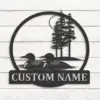 Round black metal wall sign showing two loons swimming with pine trees and moon in background, mounted on white wood planks with text panel reading "CUSTOM NAME"