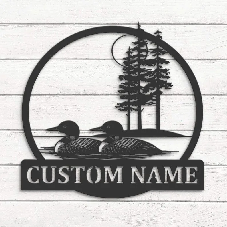 Round black metal wall sign showing two loons swimming with pine trees and moon in background, mounted on white wood planks with text panel reading "CUSTOM NAME"