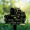 Black metal memorial sign shaped like a semi truck with text "In Loving Memory Of" and placeholder "CUSTOM NAME 1969-2023" set among daisies in a blurred green garden background