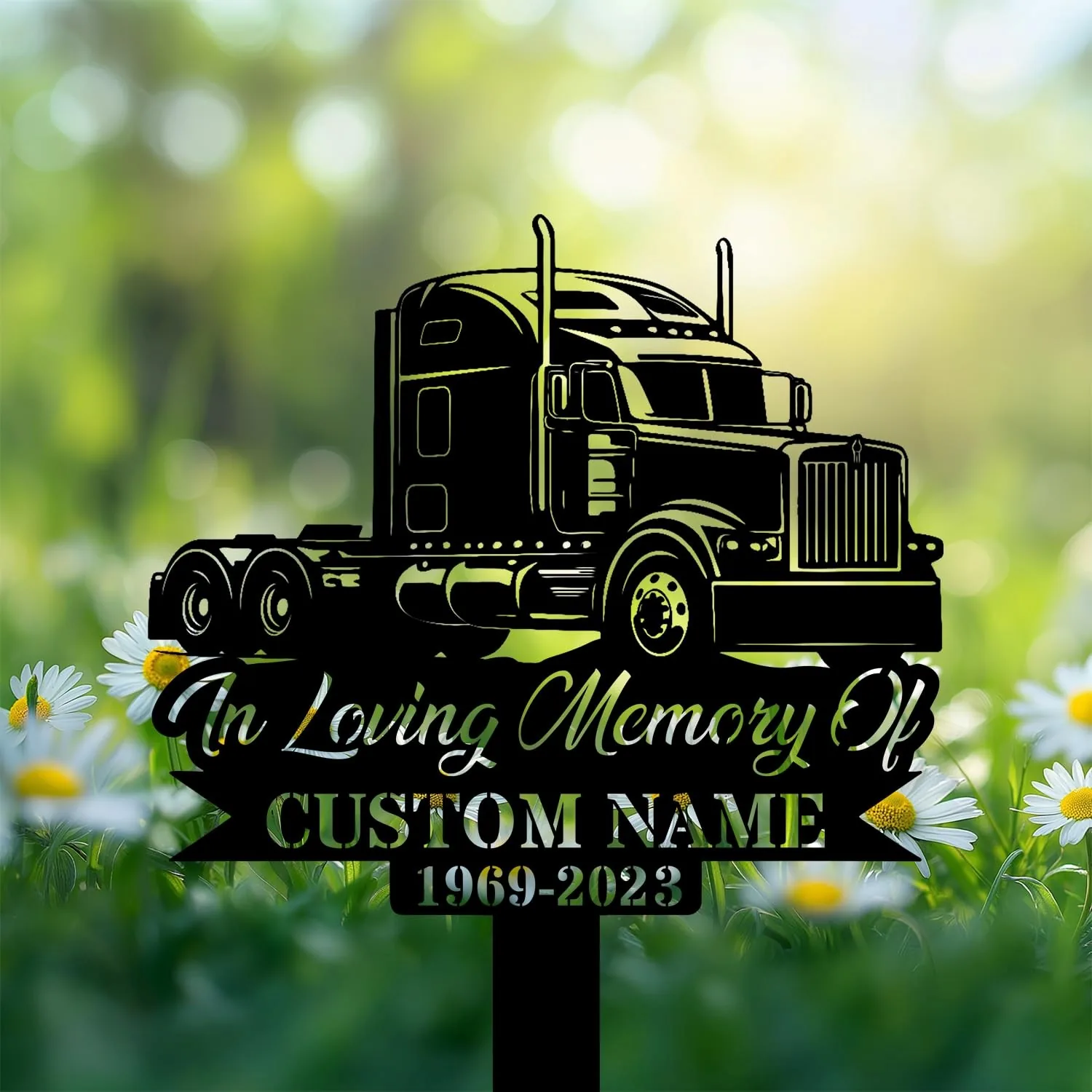 Black metal memorial sign shaped like a semi truck with text "In Loving Memory Of" and placeholder "CUSTOM NAME 1969-2023" set among daisies in a blurred green garden background