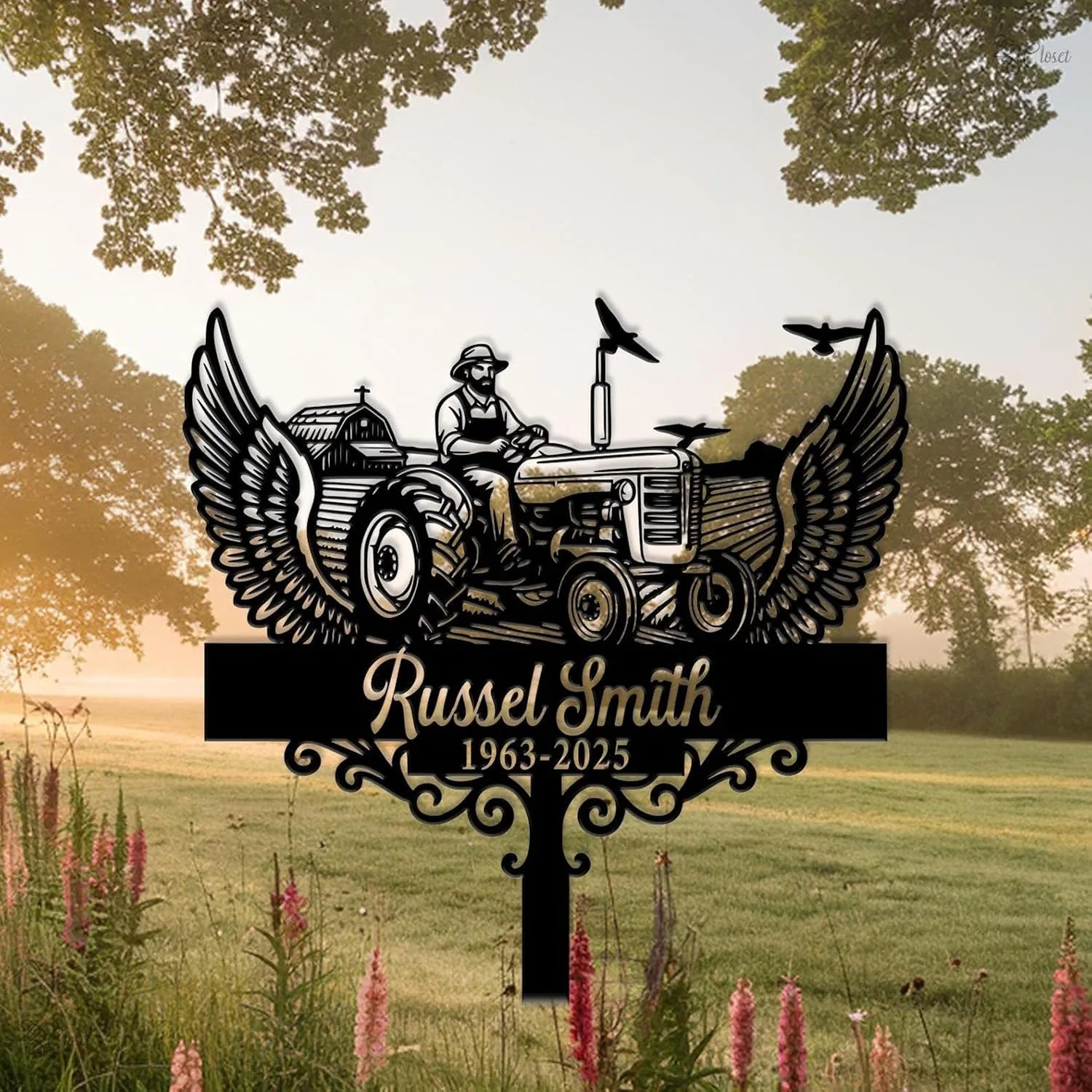 Black metal memorial yard sign showing a farmer on a tractor with wings, birds, and the name "Russel Smith" with dates "1963-2025" placed in a grassy field at sunrise
