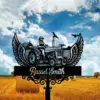 Black metal memorial sign with winged tractor silhouette and a farmer figure above a wheat field, name plaque and dates against a cloudy blue sky