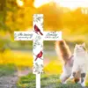 White memorial garden cross with floral pattern and red cardinals reading “In loving memory of Emily 1963–2024” placed outdoors on a sunlit lawn with a fluffy cat nearby