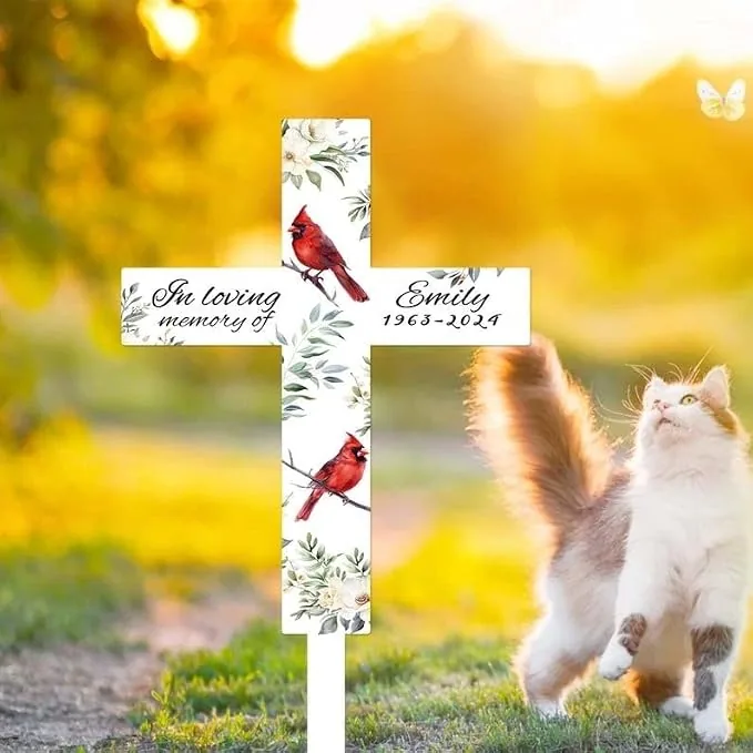 White memorial garden cross with floral pattern and red cardinals reading “In loving memory of Emily 1963–2024” placed outdoors on a sunlit lawn with a fluffy cat nearby