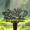 Decorative black metal lizard yard stake with floral cutouts and the name 'TILLEQUIL' and dates '2021 - 2024' against a sunlit grassy background
