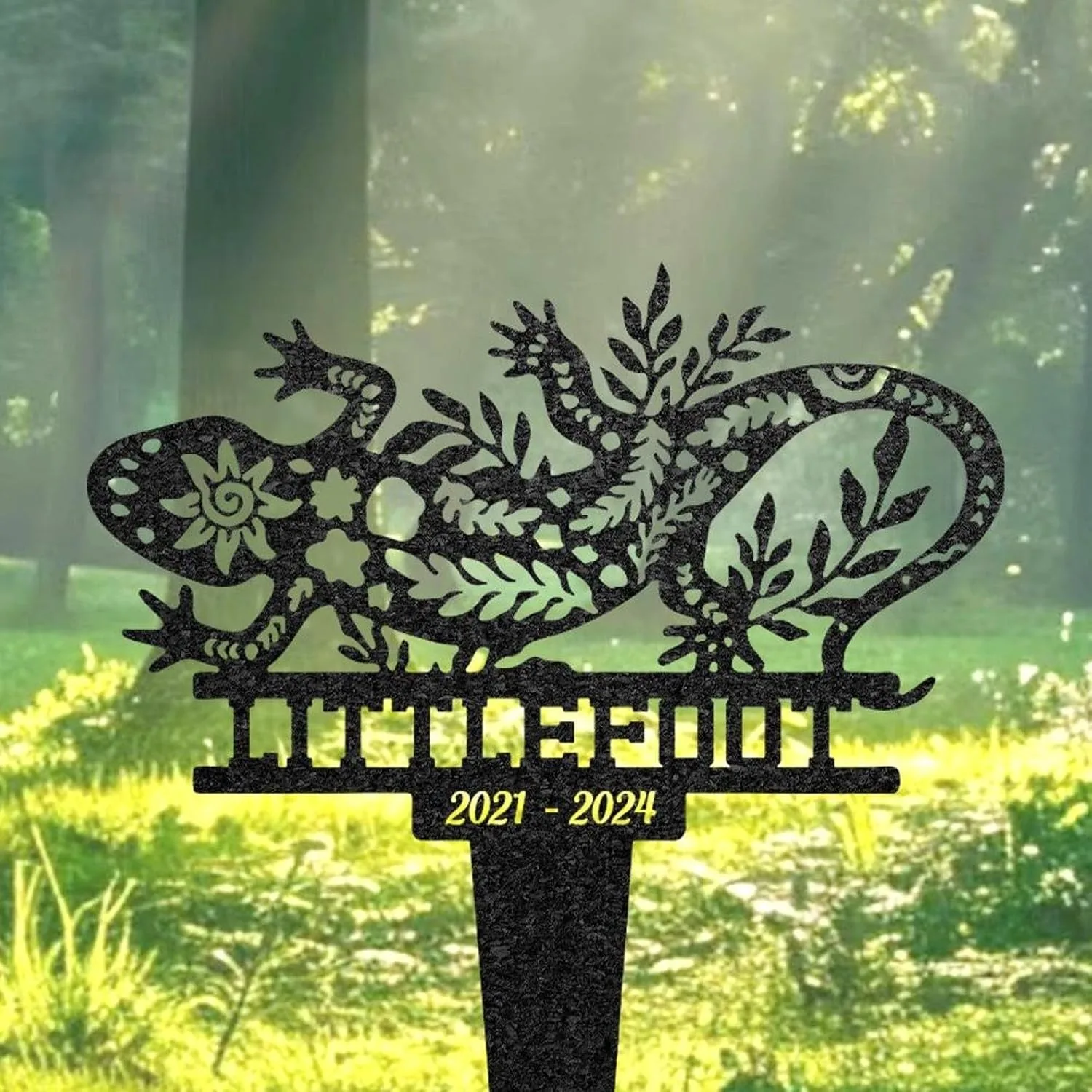 Decorative black metal lizard yard stake with floral cutouts and the name 'TILLEQUIL' and dates '2021 - 2024' against a sunlit grassy background