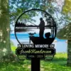 112 Black metal memorial sign reading "In Loving Memory of Jacob Henderson 1956 - 2025" with silhouette of a fisherman on a boat and a fish, set on grassy lakeside under trees
