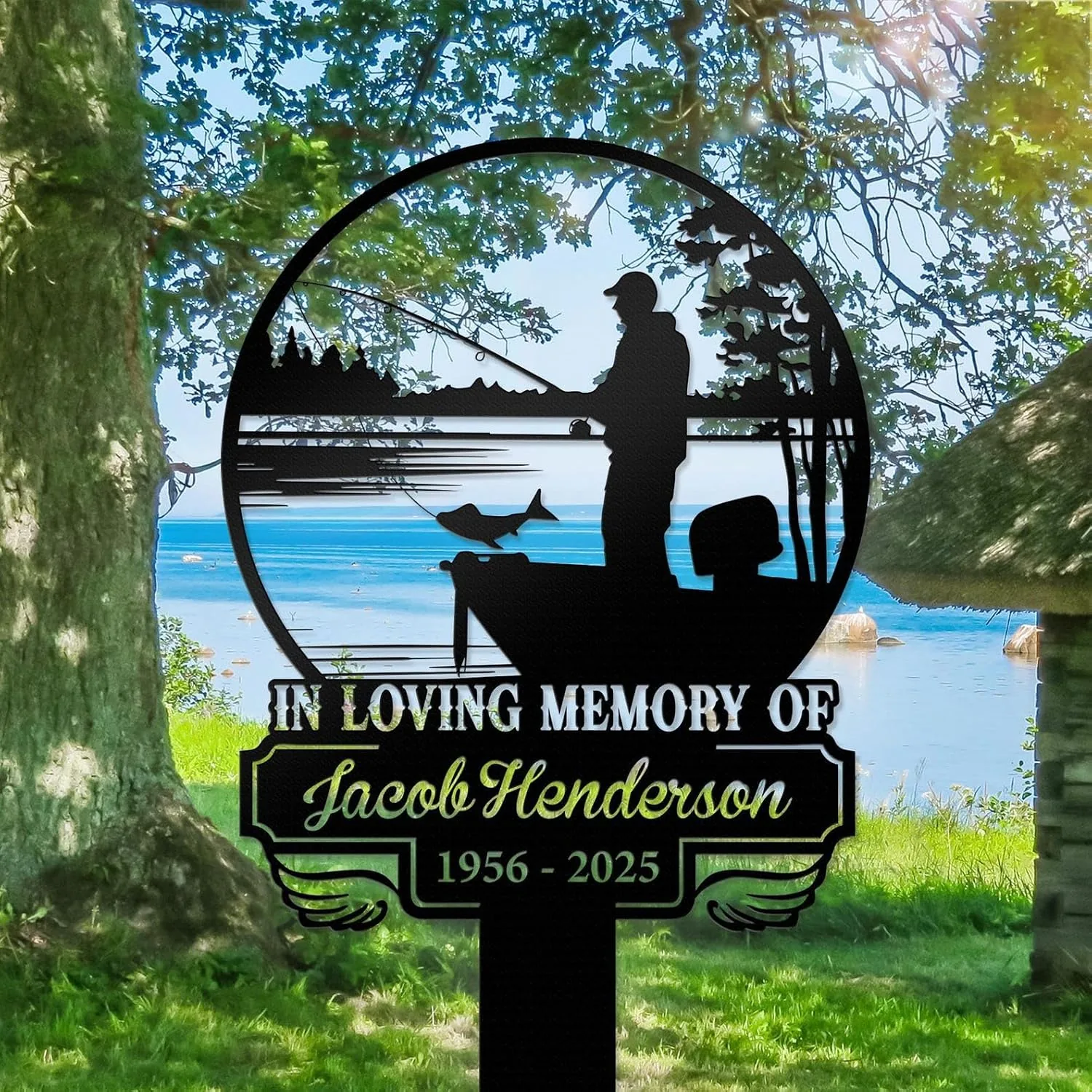 112 Black metal memorial sign reading "In Loving Memory of Jacob Henderson 1956 - 2025" with silhouette of a fisherman on a boat and a fish, set on grassy lakeside under trees