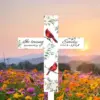 White memorial cross with floral accents and two red cardinals reading "In loving memory of Emily 1963–2024" set in a colorful wildflower field at sunset