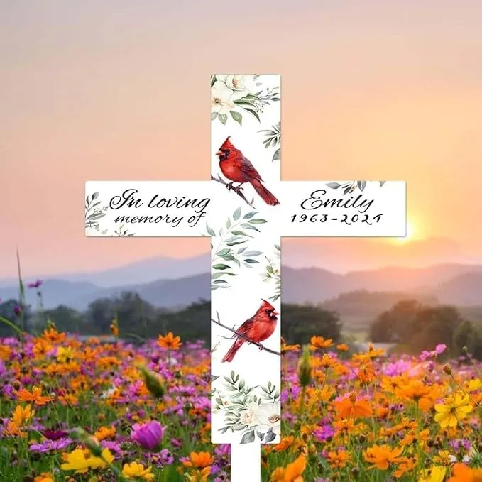 White memorial cross with floral accents and two red cardinals reading "In loving memory of Emily 1963–2024" set in a colorful wildflower field at sunset