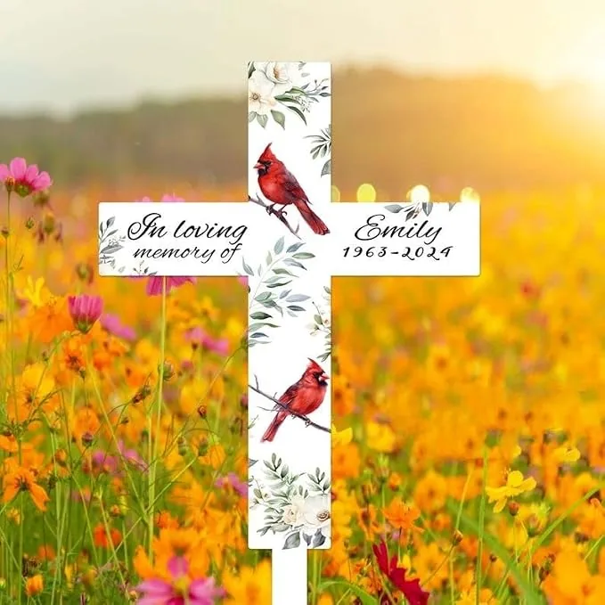 White memorial cross in a sunlit wildflower field reading "In loving memory of Emily 1963-2024" with red cardinals and floral accents