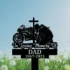 Black memorial sign with a cross, kneeling motorcyclist and bike over text reading "In Loving Memory Of DAD 1967-2025" against a blue sky and dandelion field