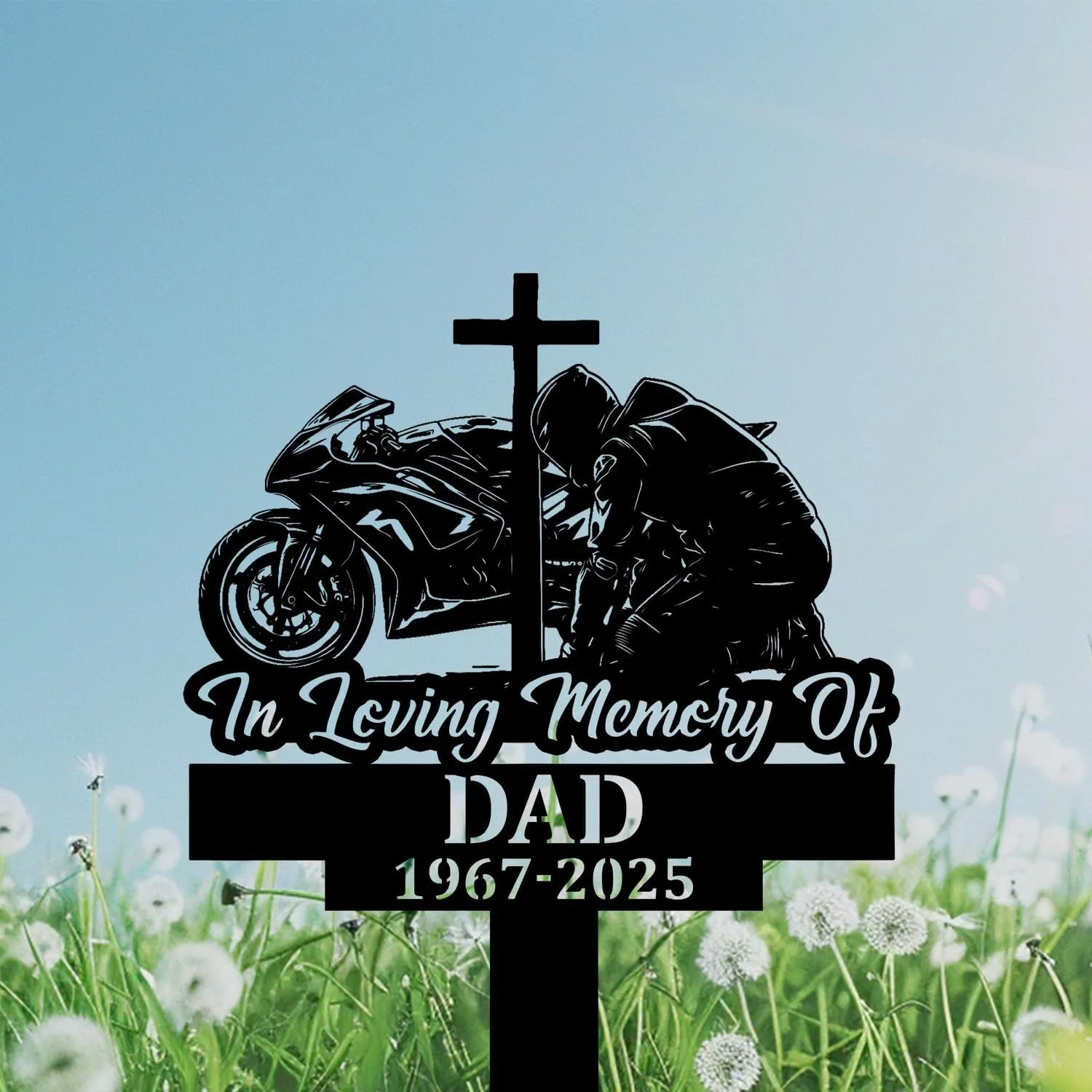 Black memorial sign with a cross, kneeling motorcyclist and bike over text reading "In Loving Memory Of DAD 1967-2025" against a blue sky and dandelion field