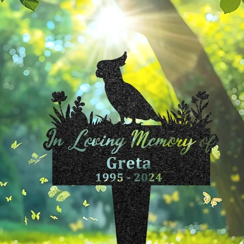 132-2 Black garden memorial stake silhouette of a cockatiel perched above flowers reading “In Loving Memory of Greta 1995 - 2024” with sunlit green trees and butterflies in background