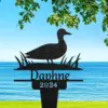 Black metal yard stake silhouette of a duck standing among reeds with the name "Daphne" and year "2024", shown against a lakeside view with blue water, grassy shore and tree branches overhead