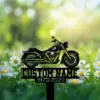 Black metal motorcycle grave marker silhouette with placeholder text "CUSTOM NAME" and years "1970-2022" placed among daisies in a sunlit green meadow