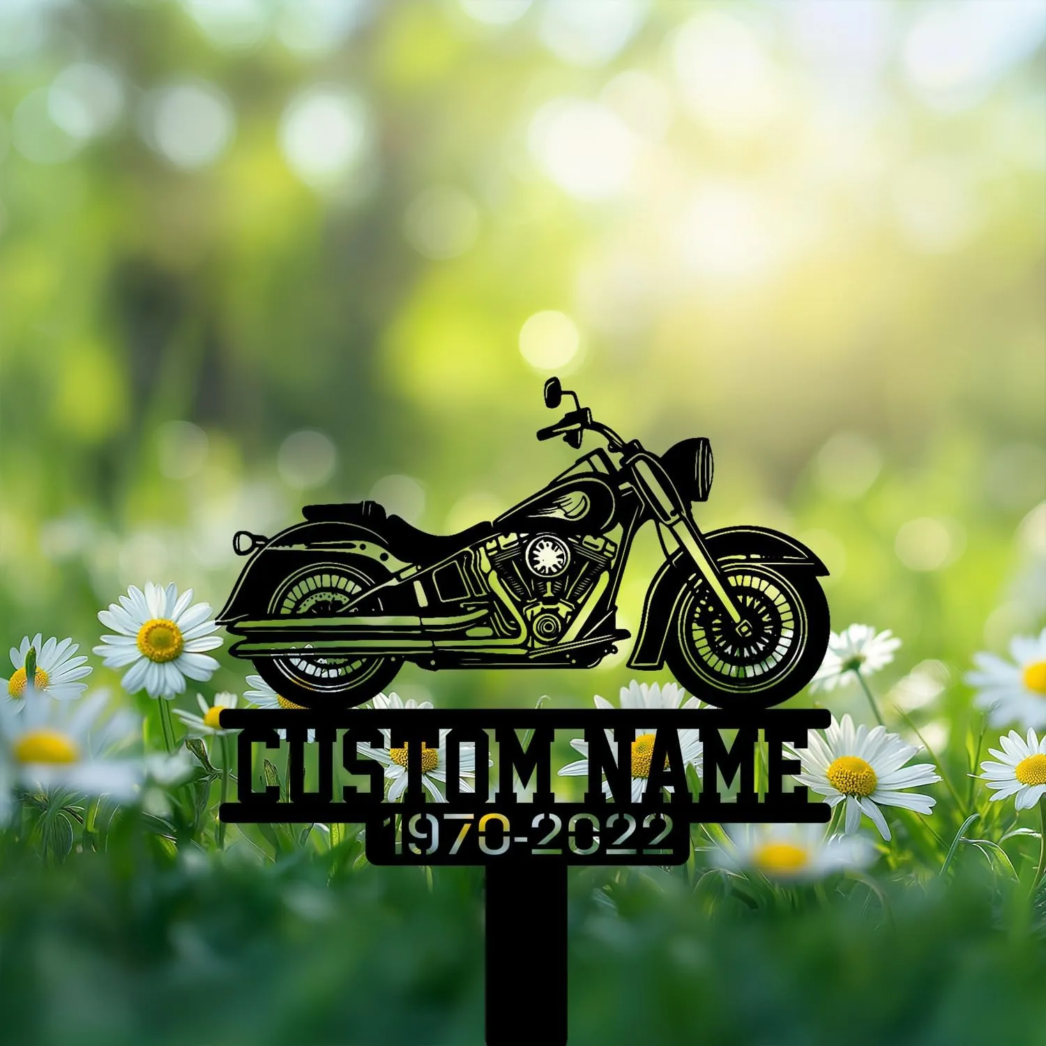 Black metal motorcycle grave marker silhouette with placeholder text "CUSTOM NAME" and years "1970-2022" placed among daisies in a sunlit green meadow
