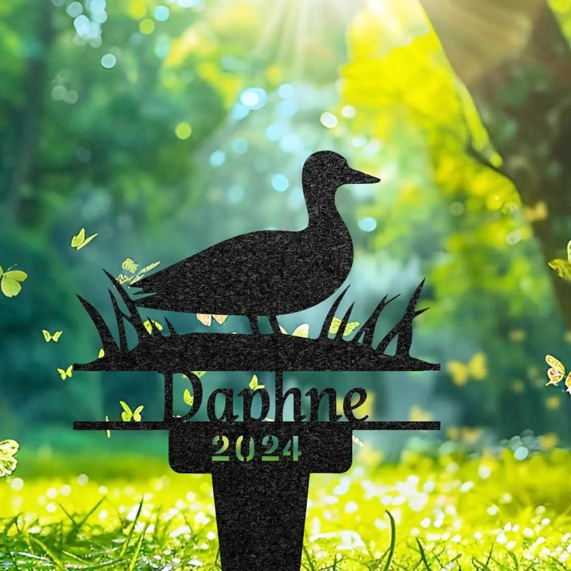 Black garden stake silhouette of a duck standing in grass with the name “Daphne” and year “2024” against a sunlit green backyard background