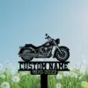 Black metal motorcycle silhouette sign with text placeholder "CUSTOM NAME" and dates "1970-2022" mounted above a grassy field of dandelions under a blue sky