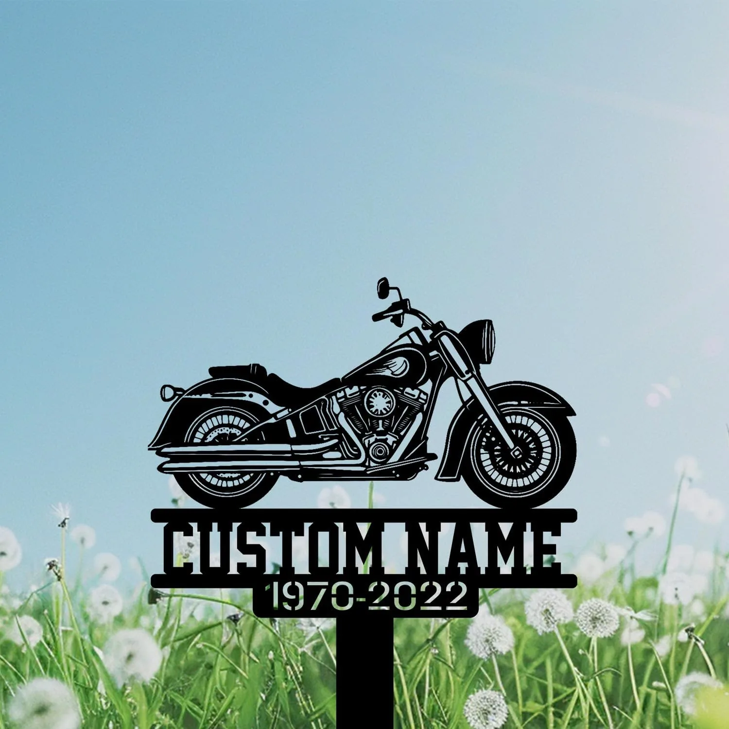 Black metal motorcycle silhouette sign with text placeholder "CUSTOM NAME" and dates "1970-2022" mounted above a grassy field of dandelions under a blue sky
