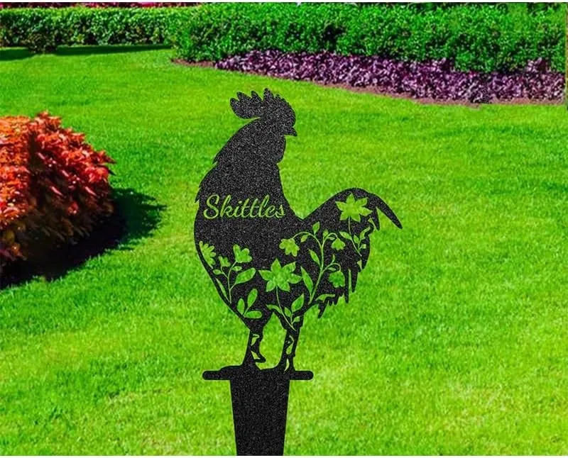 Black metal rooster garden stake with floral cutouts and the name “Skittles” planted in a green lawn near flower beds